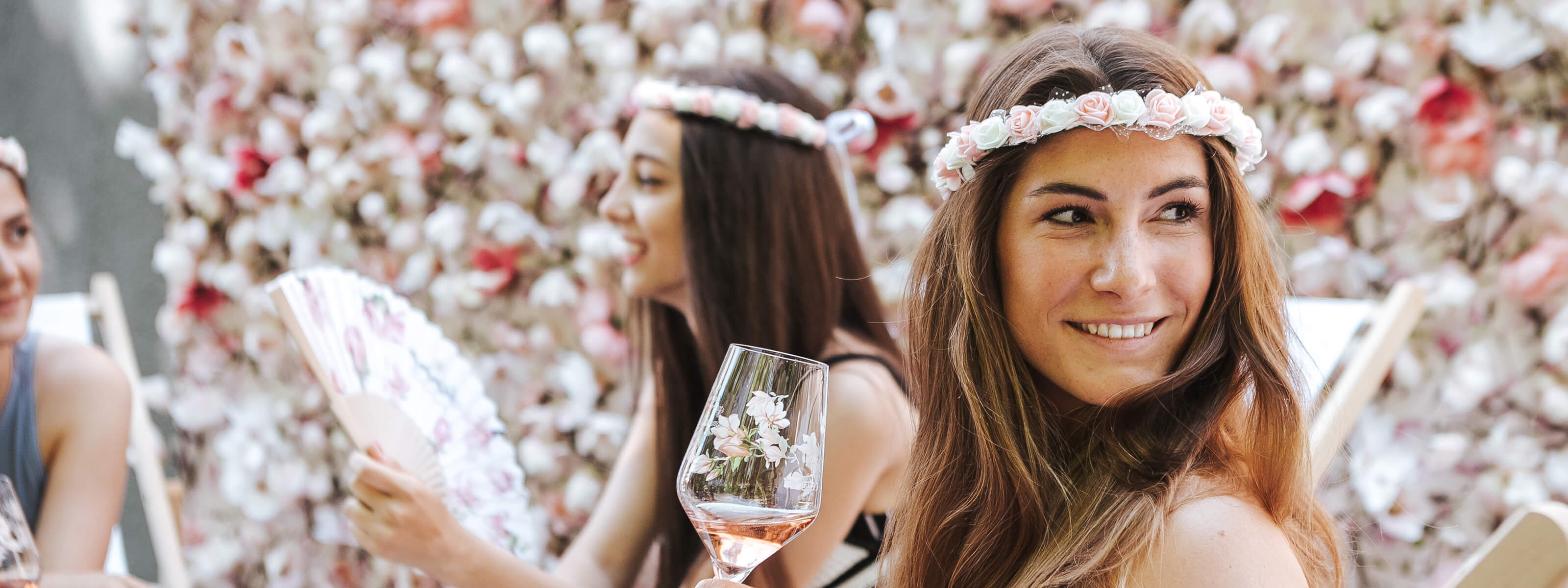 Damen sitzt mit Freunden vor einer Blumenwand mit einem Glas Sekt in der Hand