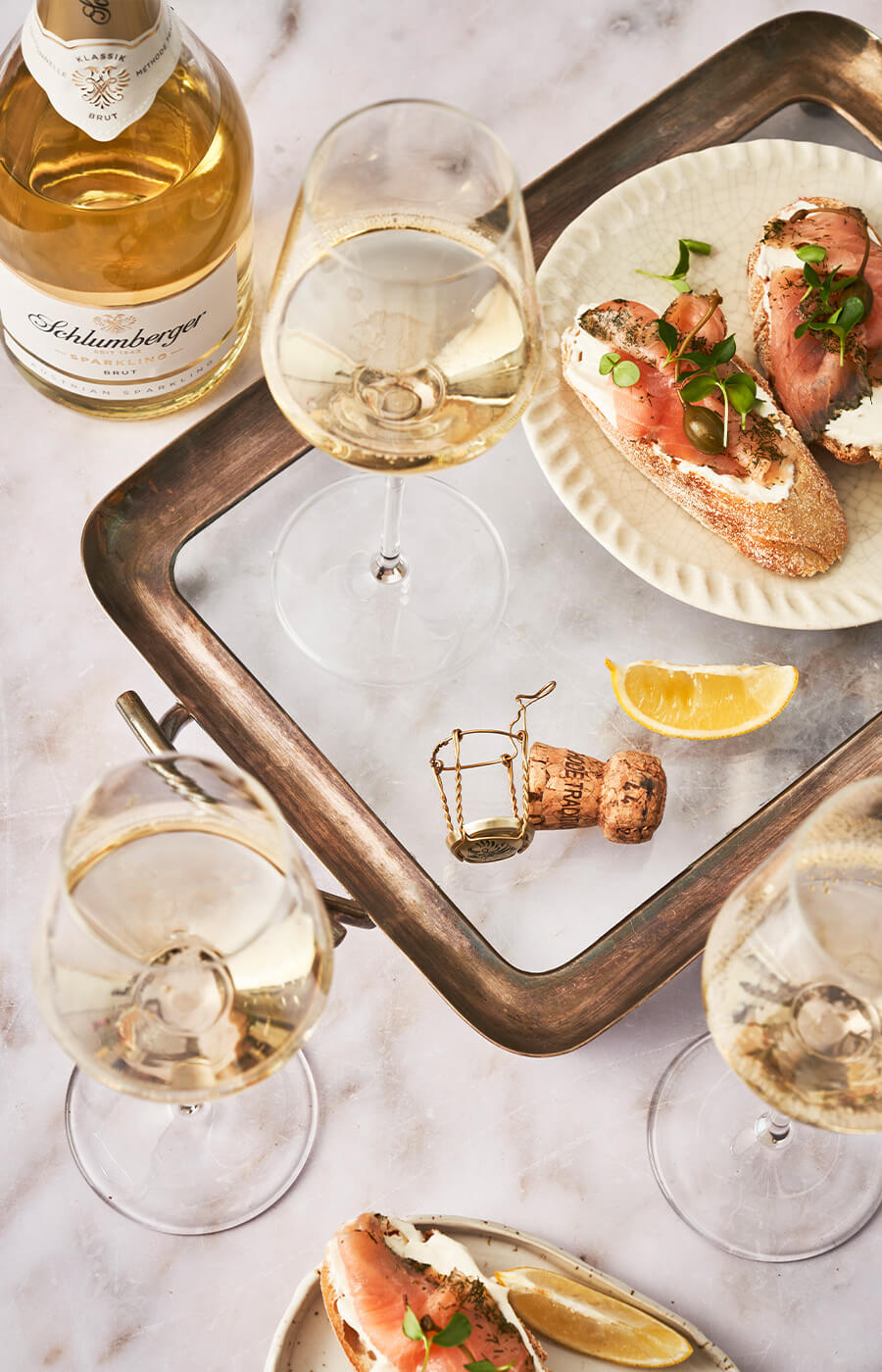 Sektgläser und eine Flasche Schlumberger Sparkling Brut auf einer marmorierten Tafel mit Lachsbrötchen.
