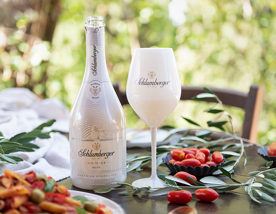 Eine geöffnete Flasche Schlumberger White Ice Secco neben einem passenden Glas, umgeben von frischen Tomaten und mediterranen Kräutern auf einem Tisch im Grünen