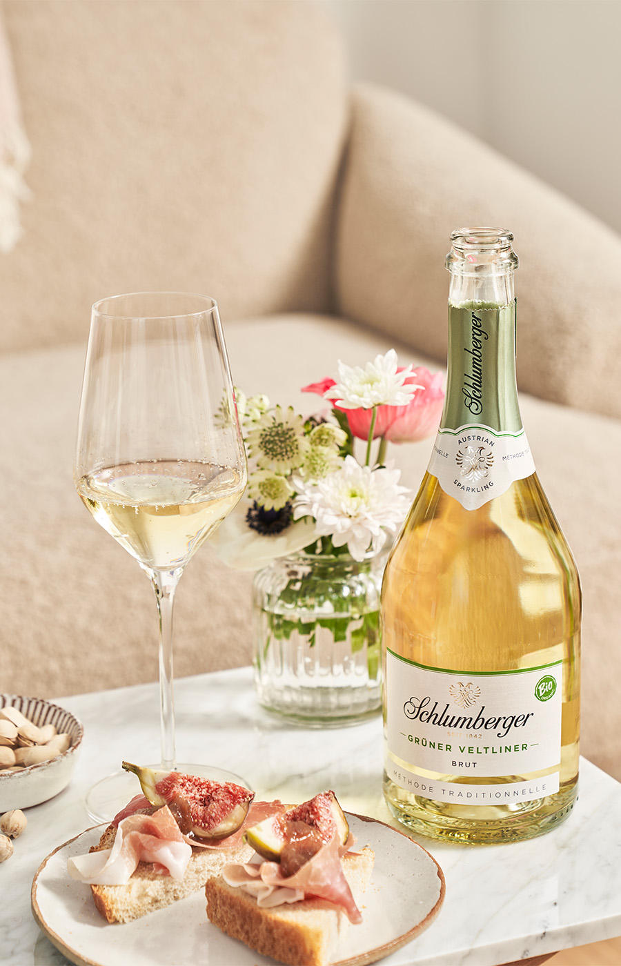 Ein Glas und eine Flasche Schlumberger Sekt auf einem Beistelltisch, gedeckt mit Blumen, Nüssen und belegten Broten