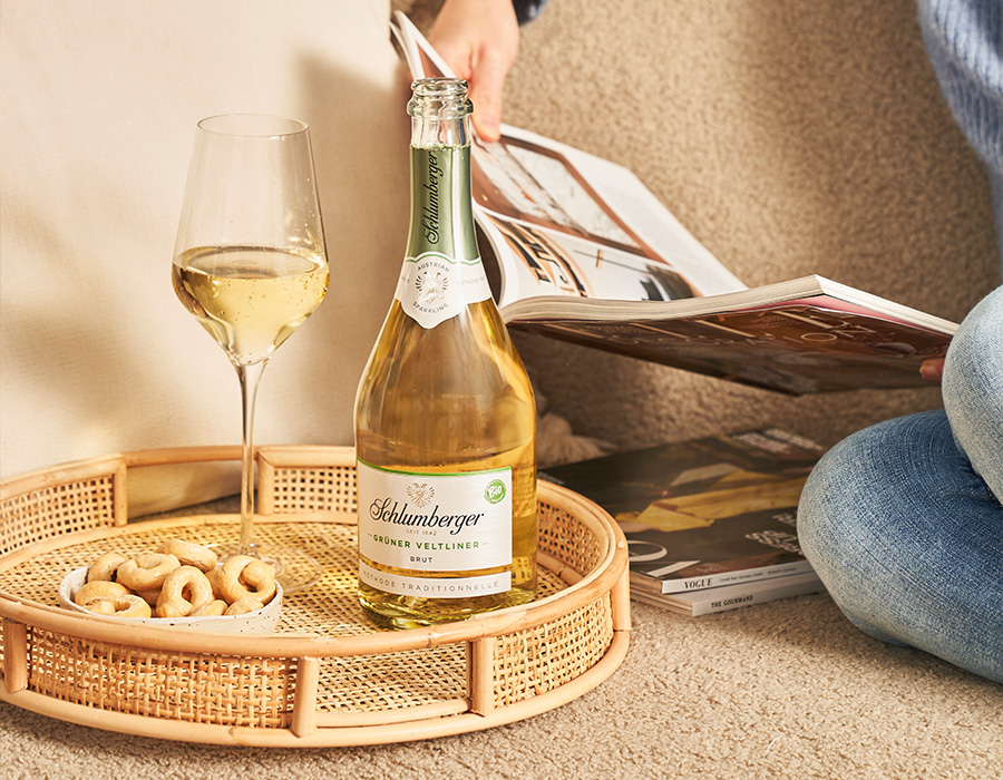 Ein Tablett mit Cashew-Nüssen, einem Sektglas und einer Flasche Schlumberger Sekt auf dem Sofa, daneben angeschnitten eine Person die liest