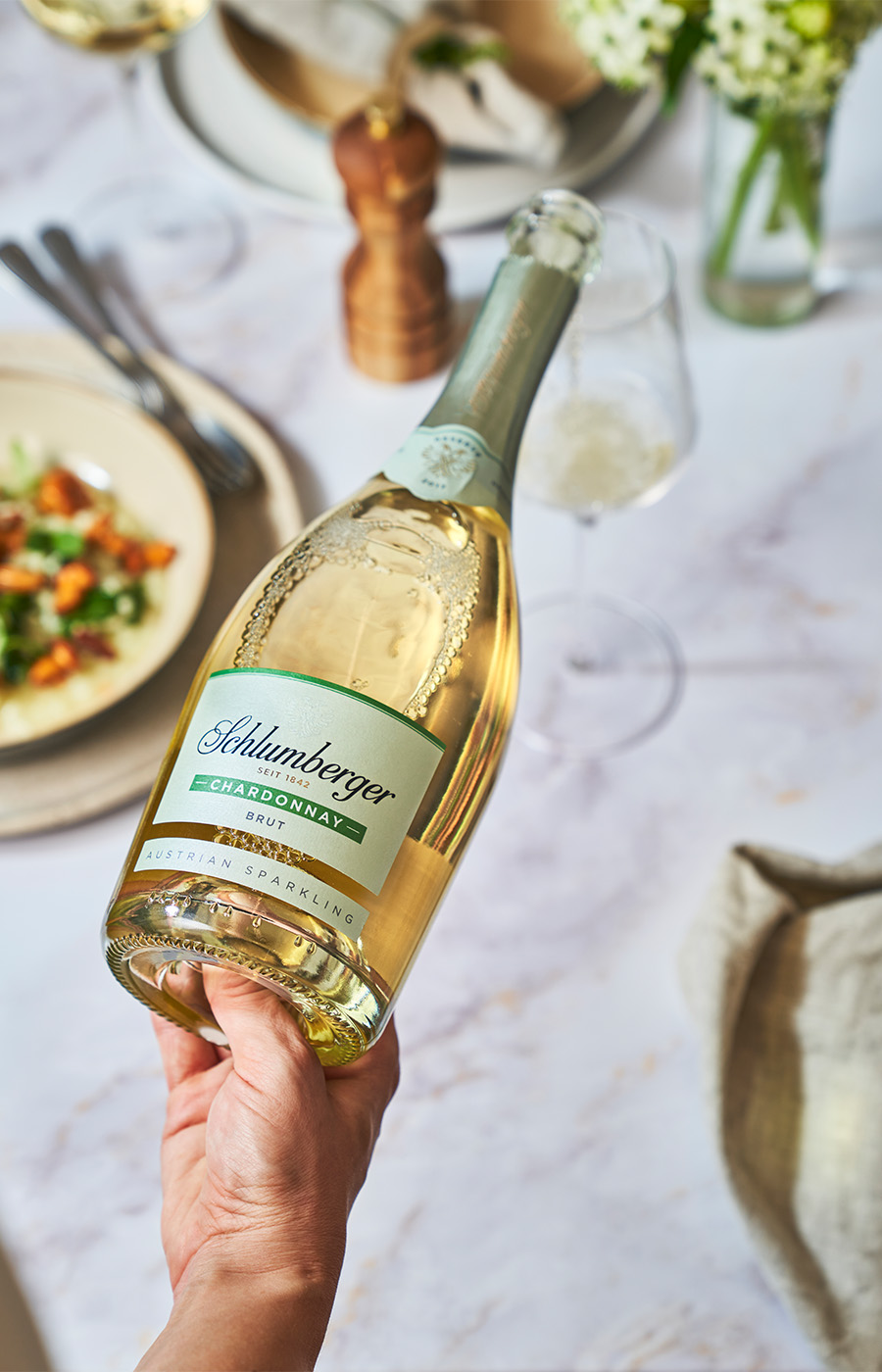 eine Hand hält eine Flasche Schlumberger Sekt, dahinter ein gedeckter Tisch