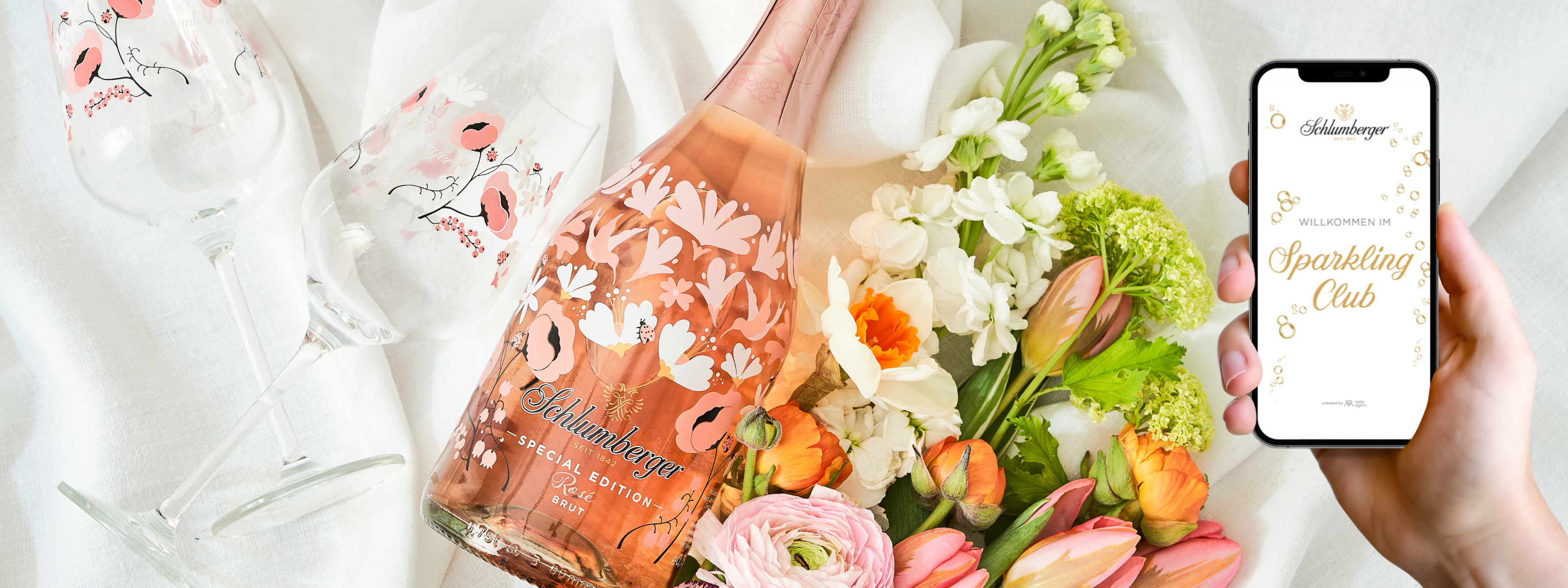 Schluberger Spring Edition, Gläser, Blumenstrauß liegen am Bett, Person hat Smartphone mit Sparkling Club App in der Hand