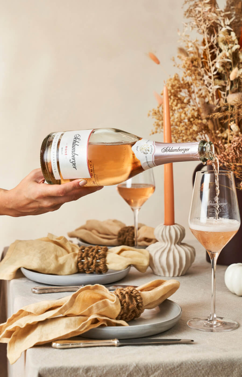 Eine Flasche Schlumberger Rosé wird in ein Glas eingeschenkt. Elegante Tischdekoration mit Servietten, Kerzen und getrockneten Blumen.