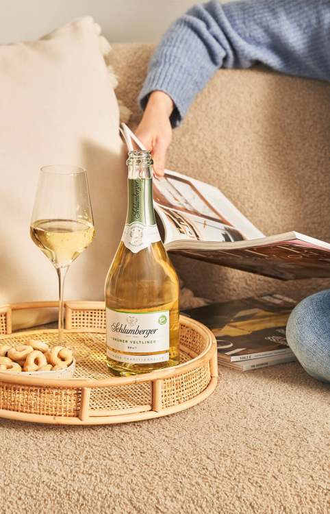 Ein Glas und eine Flasche Schlumberger Sekt auf einem Tablett, platziert auf einem Sofa, danben angeschnitten eine Person die liest