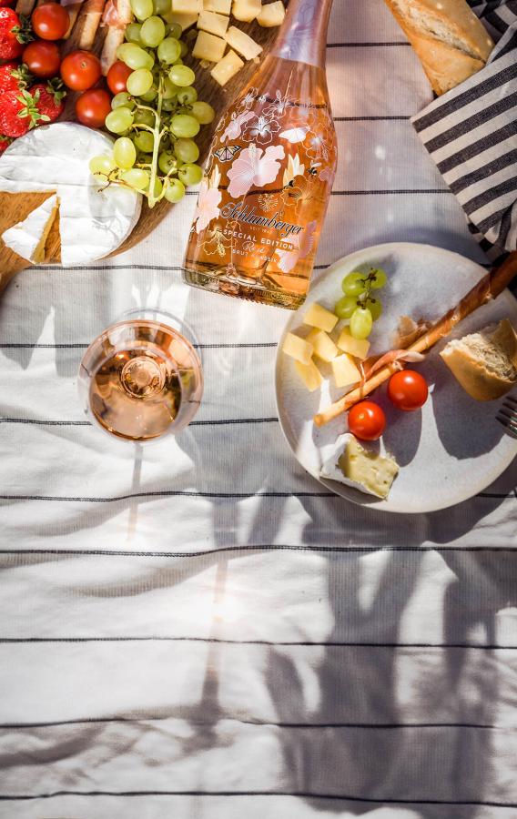 Ein sommerliches Picknick mit einer Flasche Schlumberger Sparkling Spring, einem gefüllten Sparkling Spring Glas, verschiedenen Käsesorten, Trauben, Erdbeeren, Tomaten, Baguette und Grissini, sorgfältig arrangiert auf einer gestreiften Decke unter Sonnenschein.