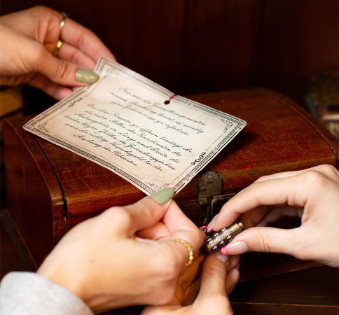 Zwei Personen öffnen ein Zahlenschloss bei einer Truhe und lesen einen Text auf einem Stück Papier