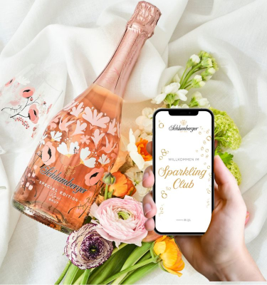 Schluberger Spring Edition, Gläser, Blumenstrauß liegen am Bett, Person hat Smartphone mit Sparkling Club App in der Hand
