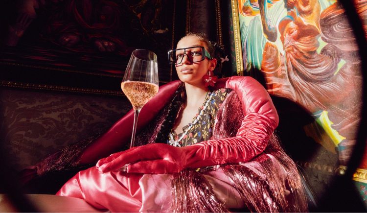 Dame in pinker Kleidung sitzt in Prunksaal mit einem Glas Rosé Secco