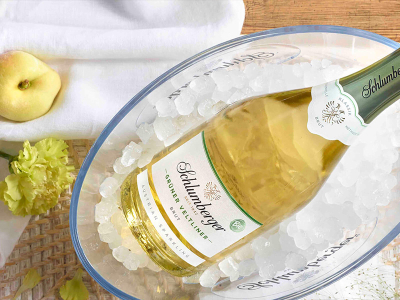 Eine Flasche Schlumberger Grüner Veltliner Brut liegt gekühlt auf Eis in einer transparenten Schale, umgeben von Pfirsich und Blumen.